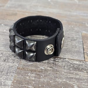 May include: A black leather bracelet with silver pyramid-shaped studs. The bracelet has a snap closure.