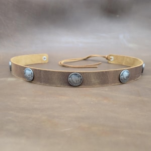 Könnte beinhalten: Ein braunes Lederstirnband mit silberfarbenen Büffelnickel-Akzenten. Das Stirnband ist mit einer Lederschnur verstellbar und hat ein Used-Finish. Das Accessoire ist für Kopfbedeckungen gedacht.