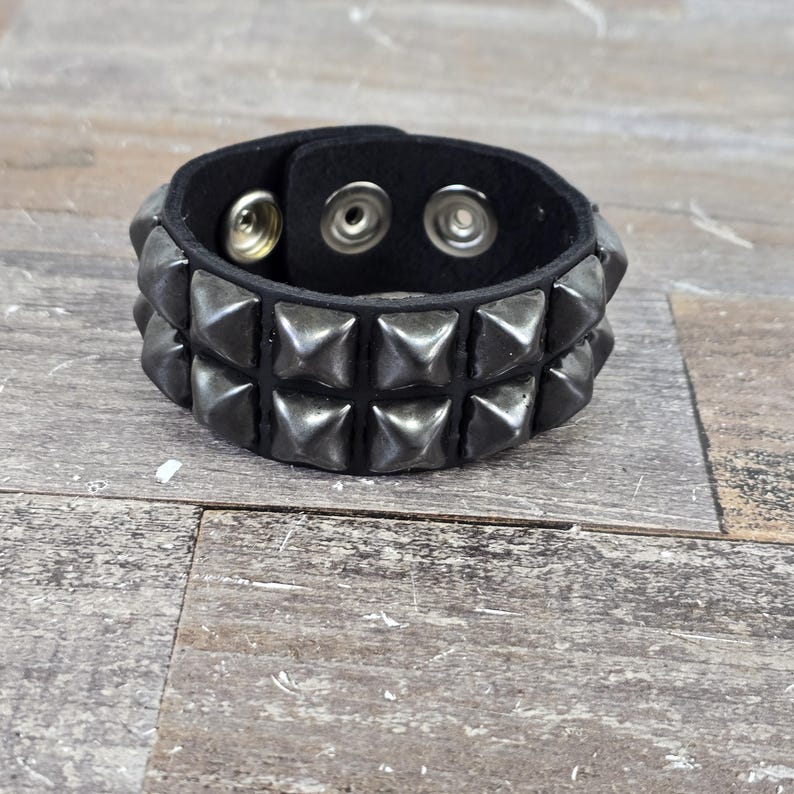 May include: A black leather bracelet with two rows of silver pyramid-shaped studs. The bracelet has three snap closures.