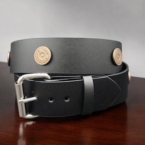 May include: Black leather belt with silver buckle and brass-colored shell casings as embellishments. The belt has a classic design with a single belt loop and a pointed end. The shell casings are stamped with the text "12 GA SHOTGUN".