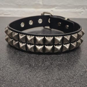 May include: Black leather collar with two rows of silver pyramid studs. The collar has a silver buckle and several silver rivets. This accessory is suitable for pets or as a fashion statement.