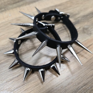 Könnte beinhalten: Zwei schwarze Lederarmbänder mit großen, silberfarbenen Metallspitzen. Die Armbänder sind geschnallt und scheinen aus einem robusten Material gefertigt zu sein. Die Spitzen sind gleichmäßig um die Bänder angeordnet und erzeugen einen kühnen, kantigen Look.