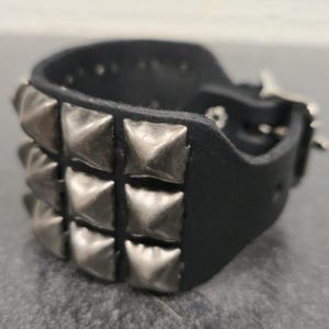 May include: Black leather cuff bracelet with rows of silver-colored pyramid studs. The bracelet has a buckle closure and is designed to be worn around the wrist. The cuff is a bold accessory.