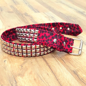 Puede incluir: Un cinturón de terciopelo rojo y negro con estampado de leopardo y tachuelas piramidales plateadas. El cinturón tiene una hebilla plateada.