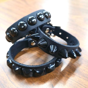 May include: Three black leather bracelets with shiny black studs and spikes. The bracelets are stacked on top of each other, showcasing the different styles of embellishments. The background is a wooden surface.