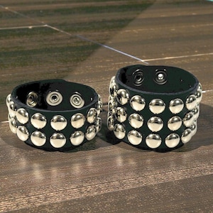 Puede incluir: Dos pulseras de cuero negro con filas de tachuelas metálicas plateadas. Cada pulsera tiene un cierre de botón a presión. Las pulseras se muestran sobre una superficie de madera marrón.