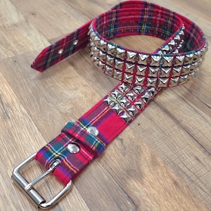 May include: A red and black plaid belt with a silver studded buckle. The belt is made of fabric and has a metal buckle with three rows of pyramid-shaped studs.