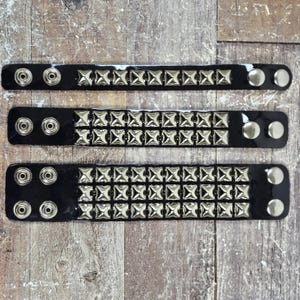 Puede incluir: Tres pulseras de cuero negro con tachuelas de pirámide plateadas. Las pulseras son de diferentes longitudes.