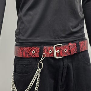 Puede incluir: Un cinturón rojo con estampado de piel de serpiente, hebilla plateada y ojales. Una cadena plateada cuelga de la trabilla del cinturón. El cinturón se lleva con pantalones negros y una camisa negra de manga larga.