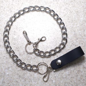 May include: A silver-colored chain with large oval links, a clasp, and a black leather strap. The chain is designed to attach to a belt loop and a wallet or keys. The hardware is silver-toned.