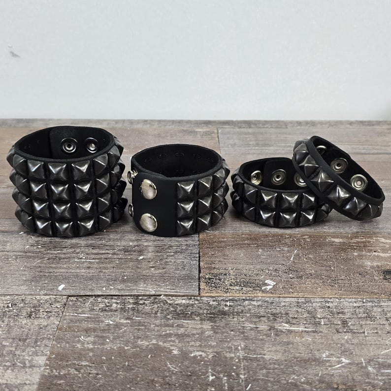 May include: Four black leather bracelets with silver pyramid studs. The bracelets are stacked on a wooden surface.