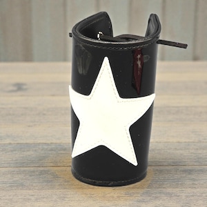 Op de afbeelding: Zwarte cilindrische accessoire met een witte sterapplicatie. Het accessoire heeft een glanzende afwerking en een kleine riem. De ster is gecentreerd op de voorkant. De achtergrond is een licht houten oppervlak.