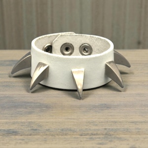 May include: White leather bracelet with silver-colored metal spikes. The bracelet has a snap closure and is designed with a punk rock aesthetic. The spikes are evenly spaced around the bracelet.