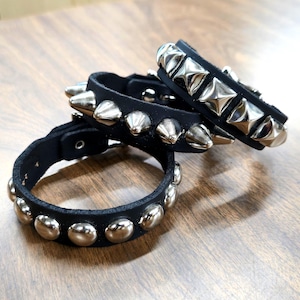 May include: Three black leather bracelets with silver metal studs. One bracelet has rounded studs, another has cone-shaped studs, and the third has square studs. The bracelets are displayed on a wooden surface.