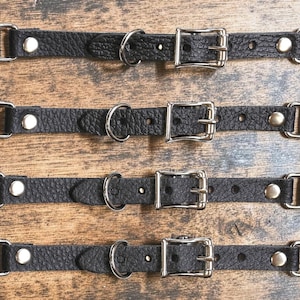 May include: Four black textured straps with silver-coloured metal hardware, including a buckle, a D-ring, and snap fasteners at each end. The straps feature a pebbled surface texture and are arranged horizontally, showcasing the adjustable design.