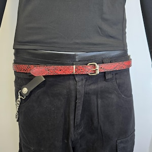 May include: A red snakeskin belt with a gold buckle and a black leather chain attached. The belt is worn around the waist of a person wearing black pants.