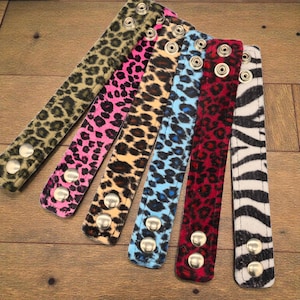 May include: A collection of six animal print bracelets in various colors, including green, pink, tan, blue, red, and black and white zebra stripes. Each bracelet has silver-toned snap closures.