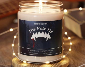 May include: A brown glass candle jar with a white label that reads "BURNING FOR The Pale Elf" with a red vampire fang graphic. The candle is lit and the jar is sitting on a wooden coaster.