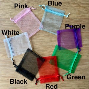 May include: A collection of seven rectangular organza drawstring pouches in pink, blue, white, purple, green, red, and black. Each sheer fabric bag features a matching ribbon drawstring closure at the top, with the color name printed near each bag.