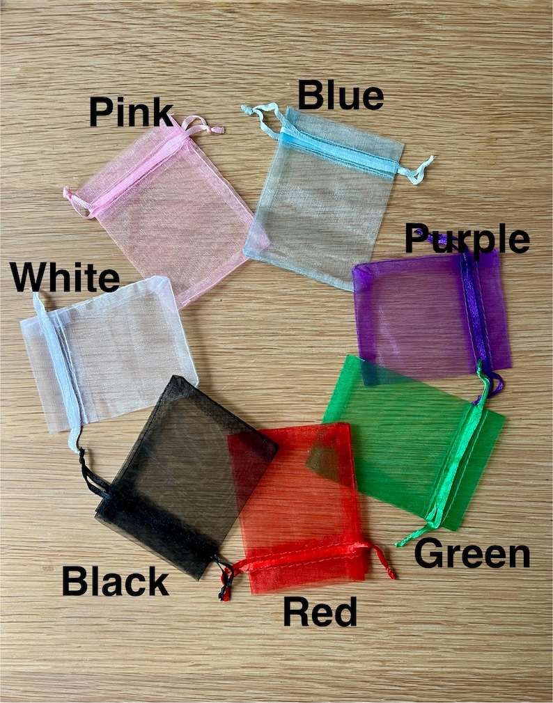 May include: A collection of seven rectangular organza drawstring pouches in pink, blue, white, purple, green, red, and black. Each sheer fabric bag features a matching ribbon drawstring closure at the top, with the color name printed near each bag.