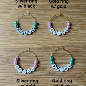 May include: Four circular hoop rings featuring white letter beads spelling BRIDE. The top left is a silver ring with teal beads, top right is a gold ring with pink beads, bottom left is a silver ring with pink beads, and bottom right is a gold ring with teal beads.