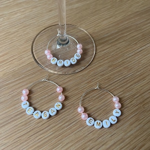 May include: Three circular silver wire wine glass charms featuring light pink beads and white alphabet beads with gold lettering. One charm spells BRIDE, one spells TEAGAN, and one spells EMILY. The charms are designed to hook onto glass stems.