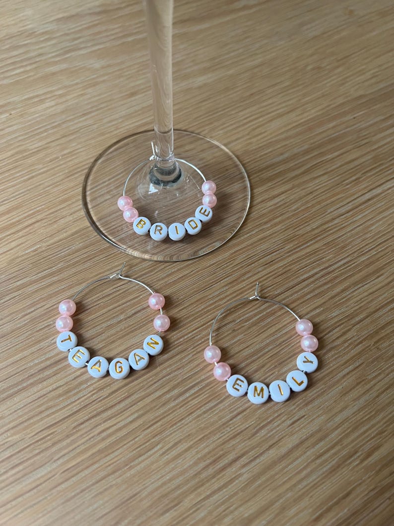 May include: Three circular silver wire wine glass charms featuring light pink beads and white alphabet beads with gold lettering. One charm spells BRIDE, one spells TEAGAN, and one spells EMILY. The charms are designed to hook onto glass stems.