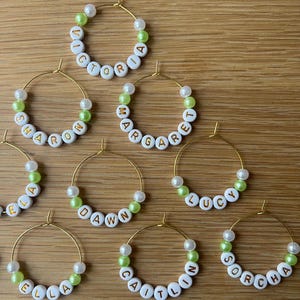 May include: A collection of gold-toned hoop earrings, each featuring white alphabet beads spelling names like Victoria, Sharon, Margaret, Dawn, Lucy, Ella, Caitlin, and Sorcha, interspersed with small white and lime green spherical beads.