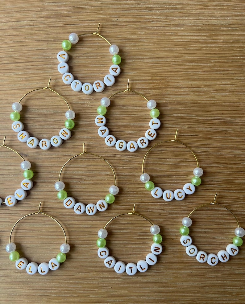 May include: A collection of gold-toned hoop earrings, each featuring white alphabet beads spelling names like Victoria, Sharon, Margaret, Dawn, Lucy, Ella, Caitlin, and Sorcha, interspersed with small white and lime green spherical beads.