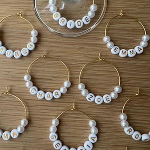 May include: A collection of gold-toned metal wine glass charms featuring white spherical beads and white circular beads with gold lettering. The charms display names including BRIDE, LAUREN, EMMA, CHAR, ZOE, MA, GEORGIA, and RO.