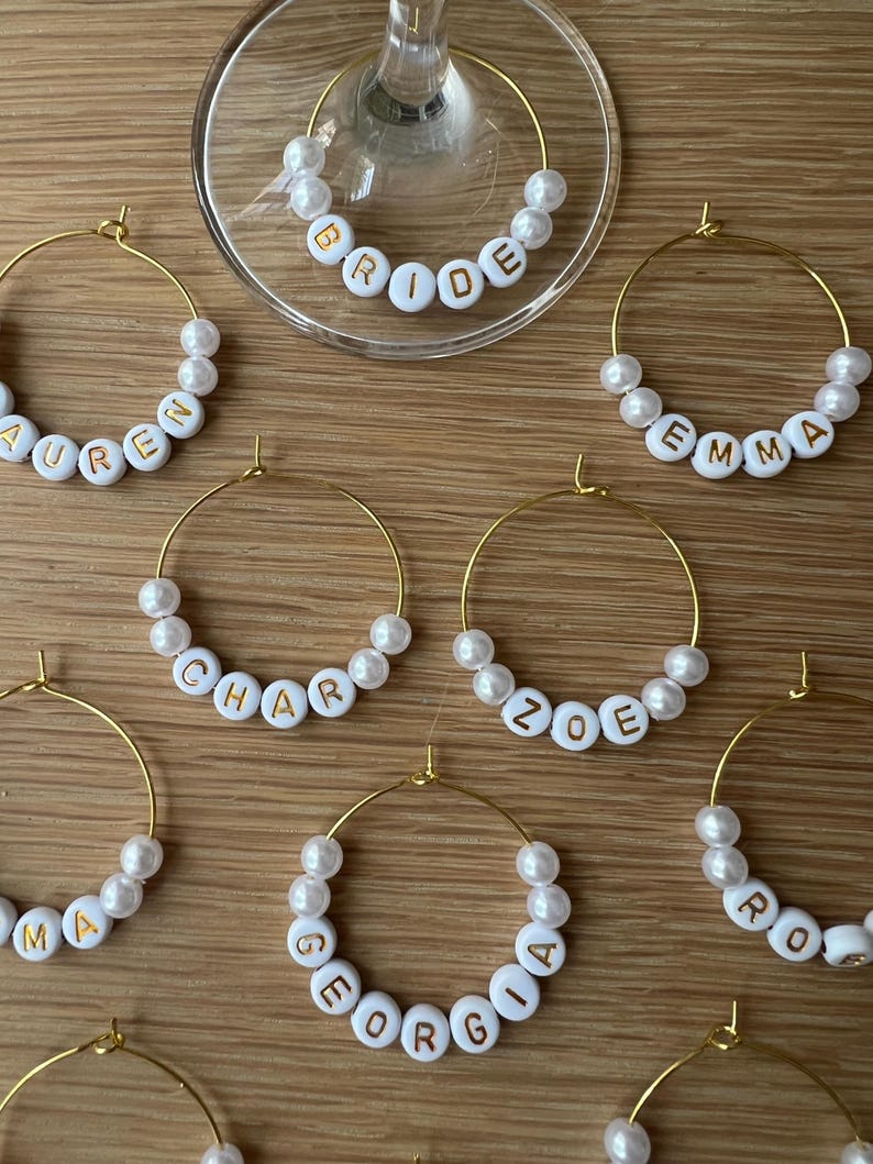 May include: A collection of gold-toned metal wine glass charms featuring white spherical beads and white circular beads with gold lettering. The charms display names including BRIDE, LAUREN, EMMA, CHAR, ZOE, MA, GEORGIA, and RO.