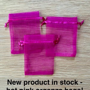 May include: Three hot pink organza drawstring bags with a sheer, semi-transparent fabric texture. The bags feature a matching ribbon closure at the top. The text "New product in stock - hot pink organza bags!" is printed in black at the bottom.