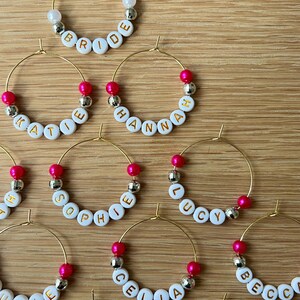 May include: A collection of gold-tone wire hoop charms featuring white alphabet beads with gold lettering spelling names like Bride, Katie, Hannah, Sophie, Lucy, Julie, Celia, and Becca, accented with hot pink and silver-tone spherical beads.