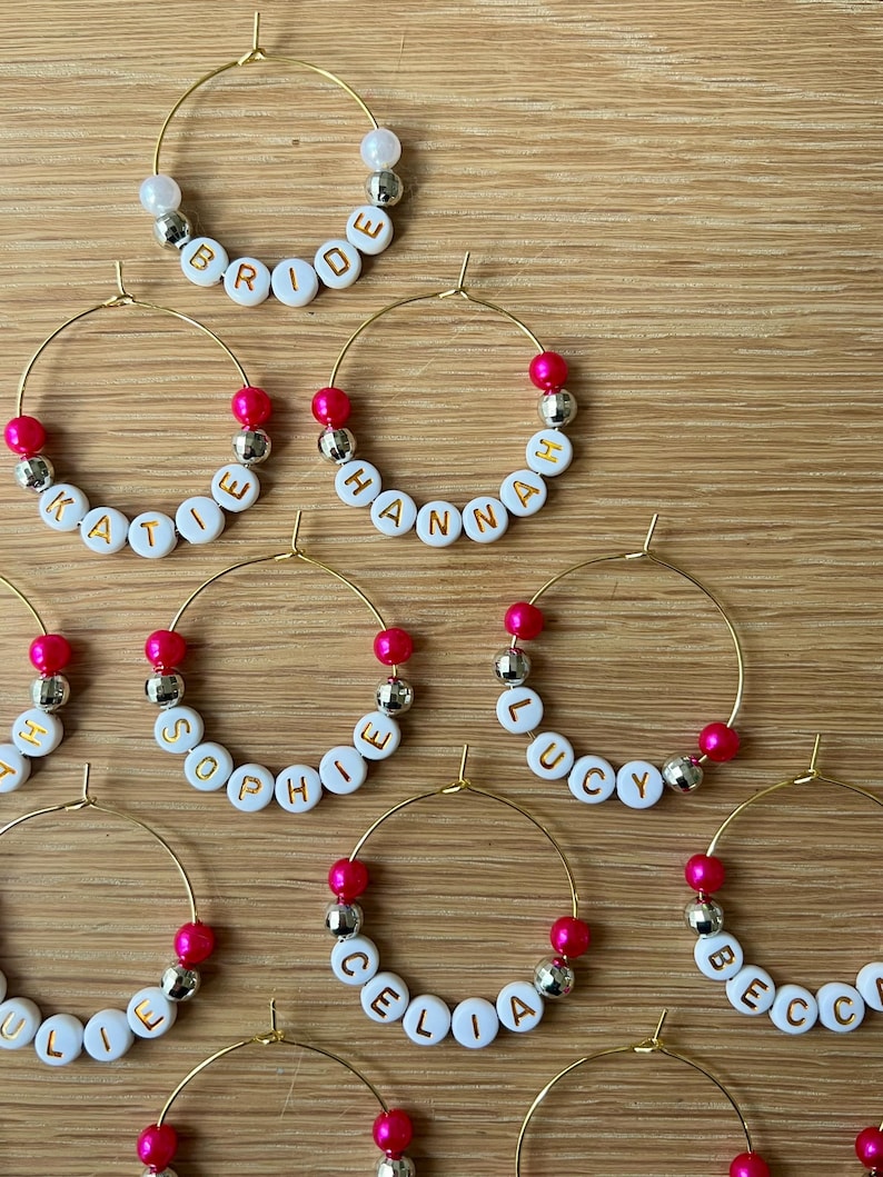 May include: A collection of gold-tone wire hoop charms featuring white alphabet beads with gold lettering spelling names like Bride, Katie, Hannah, Sophie, Lucy, Julie, Celia, and Becca, accented with hot pink and silver-tone spherical beads.
