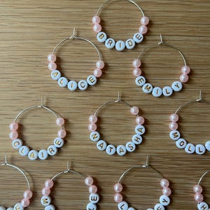 May include: A collection of silver-toned metal hoop charms, each featuring light pink beads and white circular beads with gold-colored letters spelling out names such as Bride, Alice, Emily, Claire, Natasha, Molly, Michelle, and Lesley.