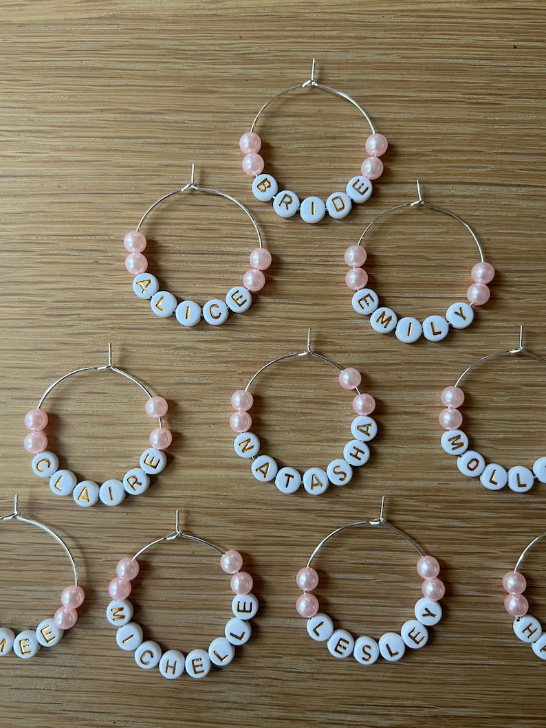 May include: A collection of silver-toned metal hoop charms, each featuring light pink beads and white circular beads with gold-colored letters spelling out names such as Bride, Alice, Emily, Claire, Natasha, Molly, Michelle, and Lesley.