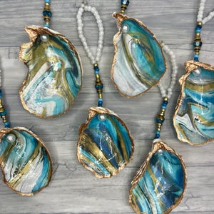 May include: Hand-painted oyster shell ornaments with gold edges, featuring swirling patterns of teal, blue, and gold. Each shell is strung with a beaded strand and a pearl accent, creating a coastal-themed decorative item.