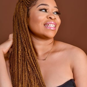 May include: A woman with long, brown braids smiles and looks to the side. She is wearing a black strapless top and a thin gold necklace.