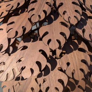 May include: Close-up of a wooden decorative piece featuring a repeating pattern of stylized Monstera leaves. The leaves are cut out of the wood, creating a play of light and shadow. The wood has a warm, natural tone.