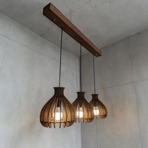 May include: Three wooden pendant lights with a warm brown finish hang from a rectangular wooden beam. Each light has a unique, open-slat design, with a bulb visible inside. The lights are suspended by black cords against a neutral gray background.