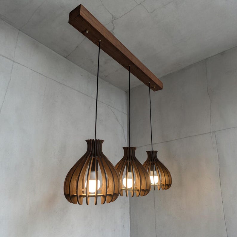 May include: Three wooden pendant lights with a warm brown finish hang from a rectangular wooden beam. Each light has a unique, open-slat design, with a bulb visible inside. The lights are suspended by black cords against a neutral gray background.