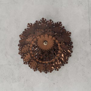 May include: A brown, circular ceiling light fixture with a leaf-like design. The light fixture is made of layered, laser-cut wood and features a central light bulb. The fixture is mounted on a gray ceiling.
