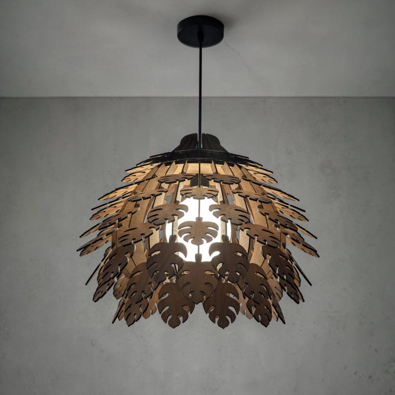 May include: A pendant light fixture with a black ceiling mount and cord. The lampshade is made of layered, laser-cut wooden leaves in a natural wood tone. The light is on, illuminating the leaf pattern.