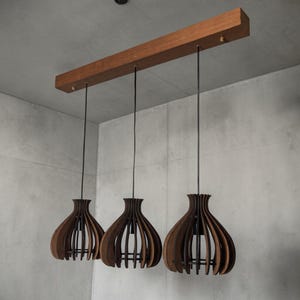 May include: A modern, three-light pendant lamp with a wooden beam and three dark brown, teardrop-shaped shades. The shades have a slatted design, and the lamp hangs from black cords. The fixture is suspended against a gray background.