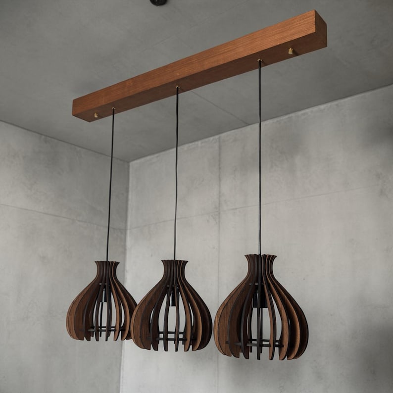 May include: A modern, three-light pendant lamp with a wooden beam and three dark brown, teardrop-shaped shades. The shades have a slatted design, and the lamp hangs from black cords. The fixture is suspended against a gray background.