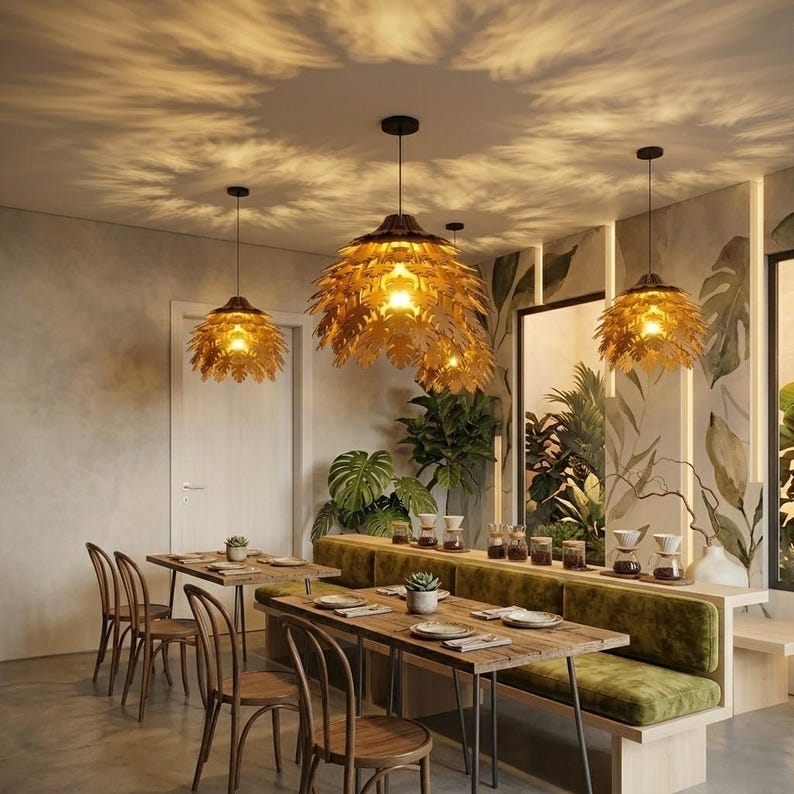 May include: A restaurant interior featuring three gold leaf-shaped pendant lights hanging from the ceiling. The lights illuminate the dining area, which includes wooden tables, chairs, and a green cushioned bench. The walls are decorated with a botanical mural.