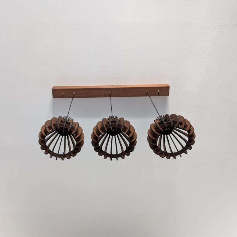 May include: A modern, three-light pendant lamp. The fixture features three circular, open-work shades made of dark brown wood, suspended from a wooden beam. The shades have a layered design with vertical slats.