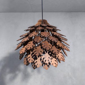 May include: A wooden pendant light fixture with a leaf-like design. The light fixture is made of multiple layers of laser-cut wood, creating a layered effect. The leaves are a warm brown color, and the fixture hangs from a black cord.