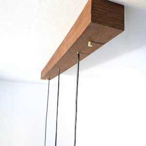 May include: A rectangular, wooden light fixture with a warm brown finish is mounted on a white ceiling. Three black cords hang down, each attached with a small, gold-colored fastener. The fixture is a modern design.