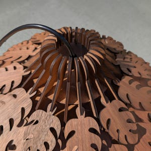 May include: A wooden pendant light fixture with a dark metal handle. The fixture is made of layered, laser-cut wood pieces in a leaf pattern. The light fixture is brown and has a circular design.